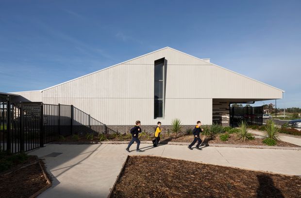 Exterior view of a contemporary single-storey building with a broad asymmetrical gabled roof, pale vertical ribbed cladding, dark brick base, narrow vertical glazing, and a covered entrance or passage. Black metal fencing, paved walkways, landscaped garden beds, and people are visible in the foreground.