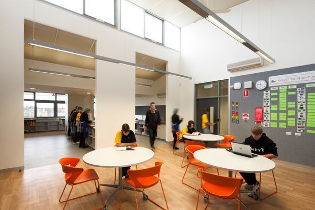 A bright, double-height communal learning or collaboration space with round white tables and vivid orange chairs. Students and staff are using the tables, with a large operational planning board, clock, pinboard, and wall-mounted air conditioner visible on the right. The room features extensive high-level glazing, exposed linear ceiling elements, pale timber flooring, and open shelving or work areas in the background.