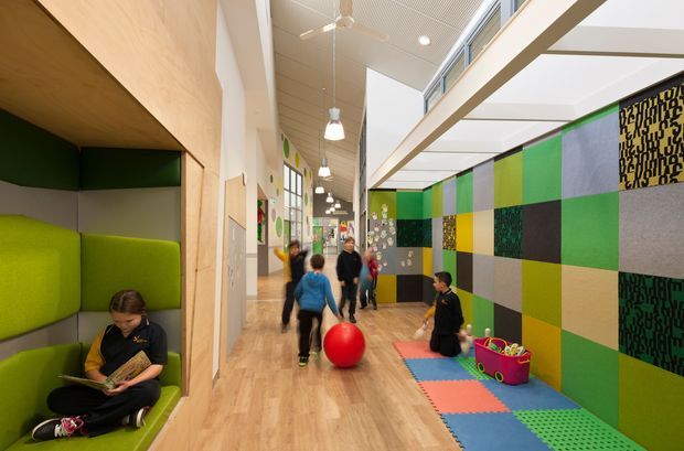 Bright, colourful multi-purpose interior corridor or activity space with padded green and grey built-in seating on the left, a graphic patchwork feature wall on the right, timber-look flooring, children and adults using the space, and a small soft-play area with colourful mats and toys. The space has a high ceiling with suspended linear lighting, ceiling fans, clerestory glazing, and an open, contemporary institutional design.