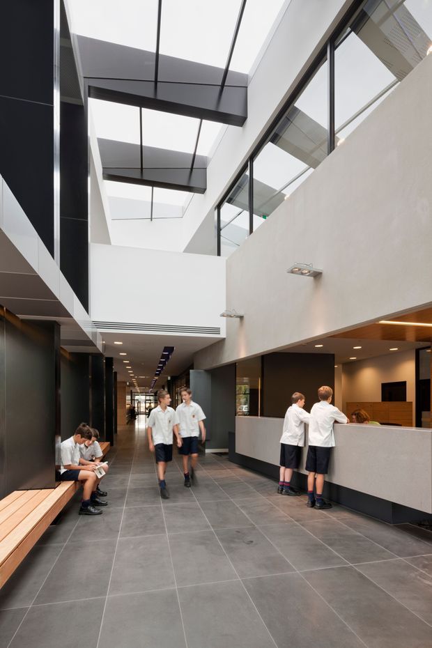 A contemporary institutional interior, likely a school reception and circulation area, with a long tiled corridor, reception counter, built-in timber bench seating, dark wall panels, and a dramatic glazed skylight atrium. Students are present, and the space is illuminated by daylight and recessed lighting.