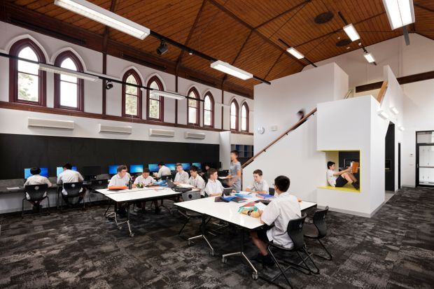 A bright, open-plan educational or commercial interior occupies a converted hall-like space. Rows of students or office users sit at grouped white tables, with a long black wall-mounted work surface at the rear. The room features a dramatic exposed timber-lined pitched ceiling with dark structural beams, a series of tall arched windows, suspended linear fluorescent lighting, dark patterned carpet, and a white stair and mezzanine enclosure with a small yellow-framed opening. The space has a contemporary adaptive-reuse character while retaining traditional architectural elements.