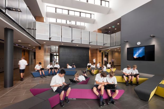 A bright, double-height contemporary communal interior resembling a school or institutional student lounge. The space features an open mezzanine with translucent-panel balustrades, clerestory glazing, exposed dark ceiling elements, timber wall accents, dark tiled flooring, and sculptural modular seating in blue, magenta, yellow, and charcoal. Several students use the lounge, with a wall-mounted display and lockers visible in the background.