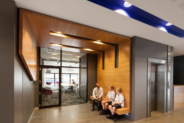 A contemporary commercial reception or lift lobby with a warm timber-lined architectural feature wall and integrated bench seating. A black-framed glazed office or meeting room is visible behind the seating, while a stainless-steel lift is set into a dark grey wall on the right. Recessed linear lighting highlights the timber surround, and large-format neutral floor tiles complete the polished interior.