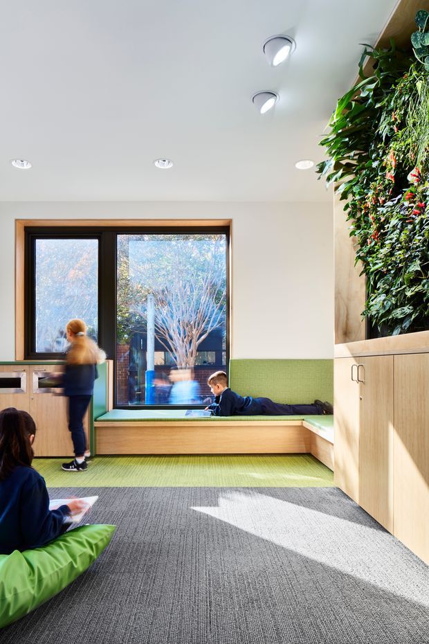 Bright, contemporary reading and waiting area with a large window, built-in timber seating upholstered in green fabric, green accent wall paneling, grey carpet, pale timber cabinetry, and a lush vertical garden feature. Several people occupy the space, which appears designed for children or a community setting.