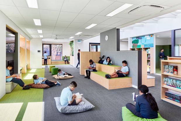 Bright contemporary school or library reading area with an open-plan layout, carpeted floor, colourful green lounge cushions, built-in timber seating and shelving, and multiple children and adults reading or relaxing. A suspended acoustic-tile ceiling with linear fluorescent lighting and exposed air-conditioning contributes to the institutional interior. Glazed openings and timber-framed partitions connect the space to adjacent rooms.