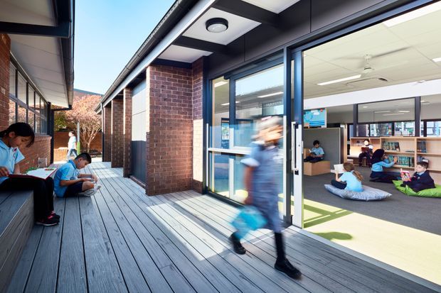 A bright school or educational facility with a covered timber-decked external walkway and brick facade. Large glazed doors open into a contemporary learning commons or classroom, where students sit on cushions and use tables and shelving. Several students are also seated along the covered walkway, and a blurred student is walking through the entrance. The building combines dark-framed glazing, red-brown brickwork, exposed outdoor decking, and a light-filled, flexible interior learning space.