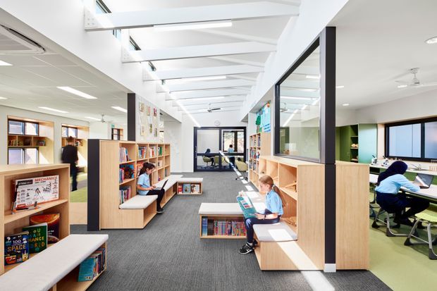 Bright contemporary school library and learning commons with open shelving, built-in timber bookcases, integrated bench seating, study tables, and adjacent classrooms visible through internal glazing. A linear skylit ceiling structure brings abundant daylight into the carpeted central circulation space, with green flooring in the study area to the right.