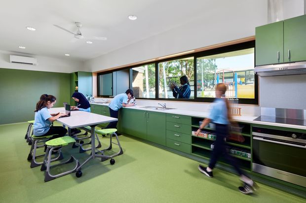 A bright, contemporary school or educational kitchen classroom with green cabinetry, green resilient flooring, white worktops, and a long run of windows above the counter. Several students are working at tables and along the kitchen benches. The room includes an induction cooktop, stainless-steel rangehood, built-in oven, sink, open shelving, ceiling fans, and recessed lighting.