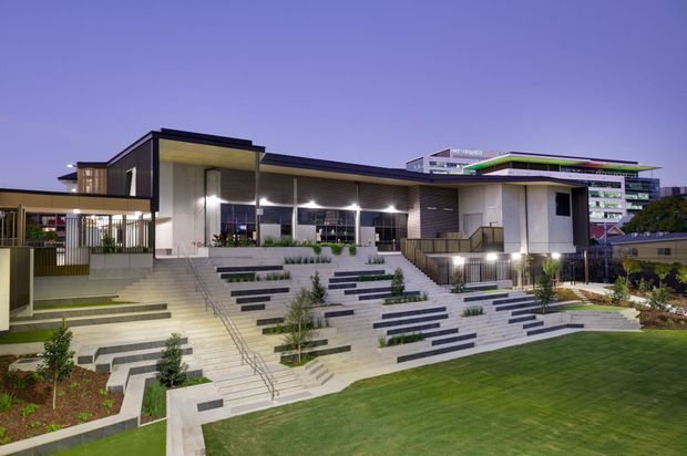 A dusk exterior view of a contemporary multi-level institutional or commercial building. The main structure features crisp geometric volumes, broad roof overhangs, extensive glazing, concrete terraces and stepped amphitheatre-like landscaping with integrated seating, planting beds and stairways. A larger multi-storey building is visible behind it, with a landscaped lawn in the foreground.