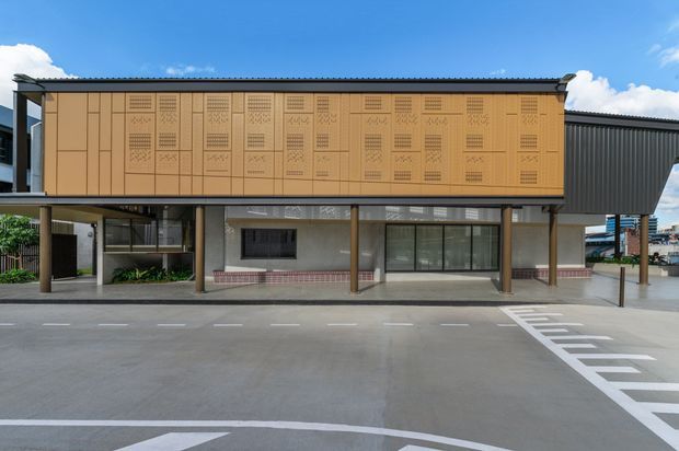 Contemporary two-storey commercial building facade with a long upper-level tan perforated-panel screen, dark metal roof and side cladding, glazed ground-floor frontage, and slender supporting columns. The building faces a broad paved forecourt with painted parking or traffic markings.