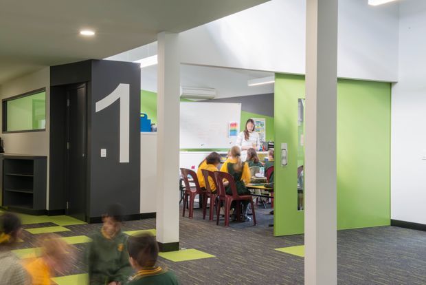 Bright contemporary educational or commercial interior showing an open circulation area leading into a classroom or activity room. The space features white structural columns and walls, vivid lime-green doors and wall panels, dark charcoal joinery and a large numeral graphic, grey patterned carpet with lime accents, and children gathered around tables in the background.