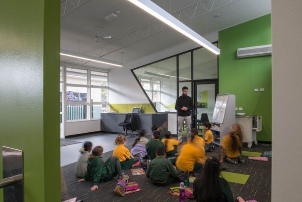 A bright contemporary classroom or learning space viewed from an open doorway, with children seated on a dark carpeted floor facing an adult presenter and a flip chart. The room has vivid green and yellow painted walls, extensive internal glazing with black frames, exposed white roof trusses, suspended linear lighting, ceiling fans, and loose classroom furniture and equipment.