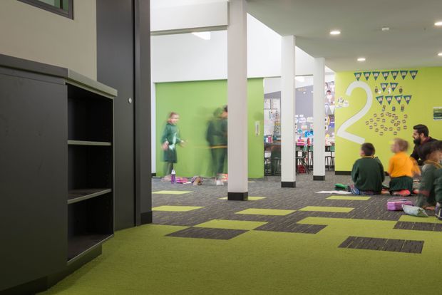 A bright contemporary educational or commercial interior with an open communal area. The space features lime-green walls and carpet, dark grey patterned carpet tiles, white structural columns, recessed ceiling lights, dark built-in shelving, and a large wall graphic with the number 2 and triangular motifs. Several children and adults are gathered in the background.