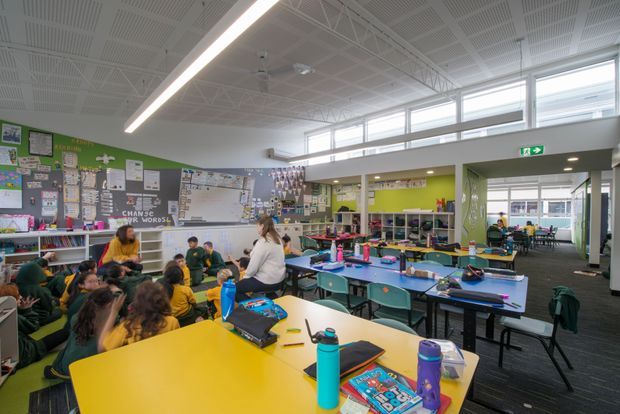 A bright, open-plan school classroom with students gathered around colourful tables and a teacher or adult facilitator. The room features extensive wall displays, open shelving, sinks and classroom storage, large clerestory-style windows, exposed white roof trusses, linear LED lighting, and a mezzanine-like internal overlook. The fit-out uses vivid yellow, teal, blue, and lime-green accents against a neutral architectural shell.