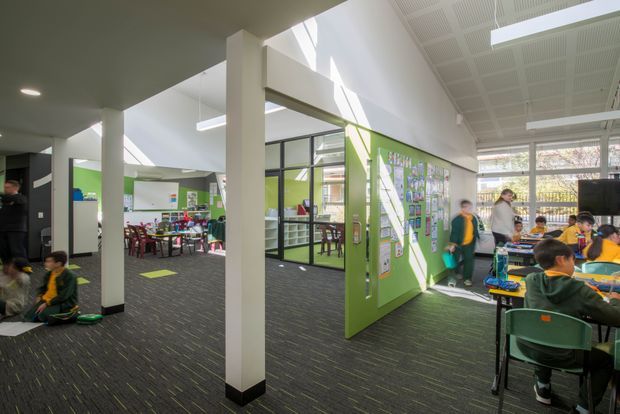 Bright contemporary school interior showing an open learning area with green feature walls, glazed classroom partitions, clustered student tables and chairs, white structural columns, acoustic ceiling panels, and large windows. The space has dark carpet flooring with scattered lime-green accents and an active educational setting.