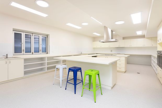 Bright, spacious contemporary kitchen with extensive white cabinetry, open shelving, pale seamless flooring, and a large central island with integrated seating. Multiple ceiling-mounted linear and recessed lights provide even illumination. The room includes stainless-steel appliances, a rangehood, a sink beneath a multi-panel window, and colourful stools in white, blue, and green.