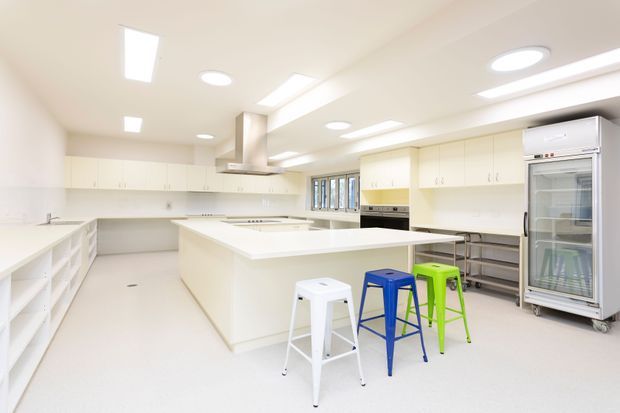 Bright, spacious commercial-style kitchen with extensive white built-in cabinetry, a large central island, open shelving, stainless-steel appliances, a commercial refrigerator, and multiple colourful metal stools. The room has a clean, functional fit-out with recessed and linear ceiling lighting and a small bank of windows.