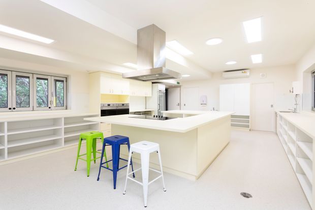 Bright, spacious contemporary commercial-style kitchen with a large central island, integrated cooking appliances, extensive white cabinetry and open shelving, multiple ceiling lights and skylights, and three colourful stools. The room has a clean, functional fit-out with pale seamless flooring and white surfaces.