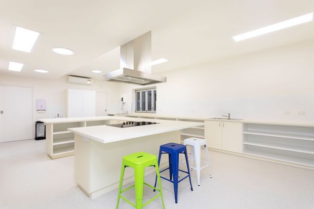 Bright, spacious all-white commercial-style kitchen with multiple long benchtops, a central island, open shelving, integrated cabinetry, stainless steel rangehood, and induction cooktop. Several colourful stools provide contrast beneath the island. The room has a clean, practical fit-out with recessed and linear ceiling lighting, a small window, and pale seamless flooring.
