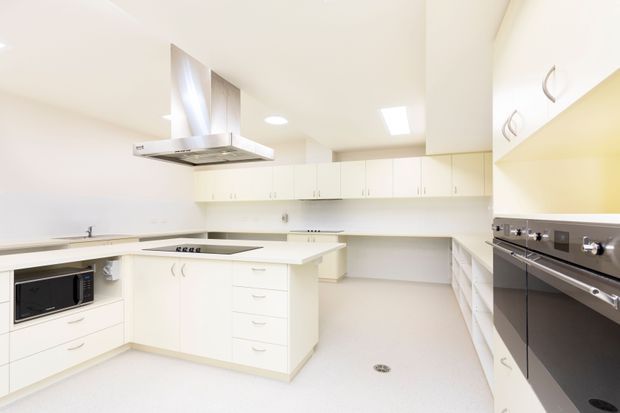 Bright, spacious white contemporary kitchen with extensive built-in cabinetry, a central island with cooktop, stainless-steel rangehood, integrated microwave, and stacked wall ovens. The room has pale seamless flooring, white benchtops and cabinetry, recessed ceiling lights, and a clean highly functional fit-out.