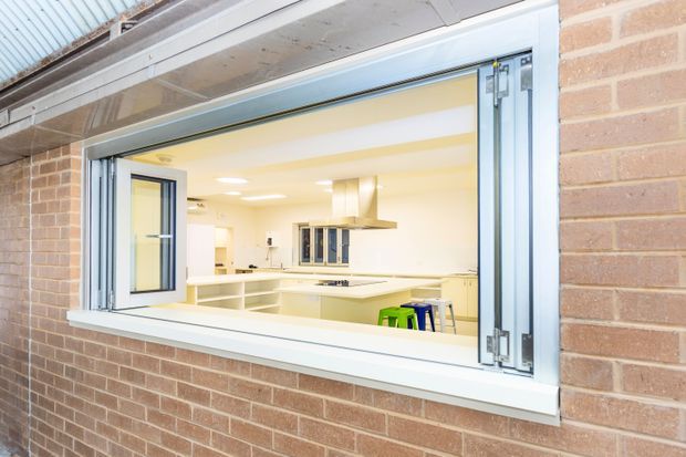 View through a wide open servery window in a red-brown brick exterior wall, looking into a bright contemporary kitchen. The opening has white-painted trim and folding or hinged aluminium-framed glazing. Inside are white cabinetry and benchtops, a central island, stainless-steel rangehood, recessed ceiling lights, and colourful stools. The image is primarily an exterior architectural view with the kitchen visible through the opening.