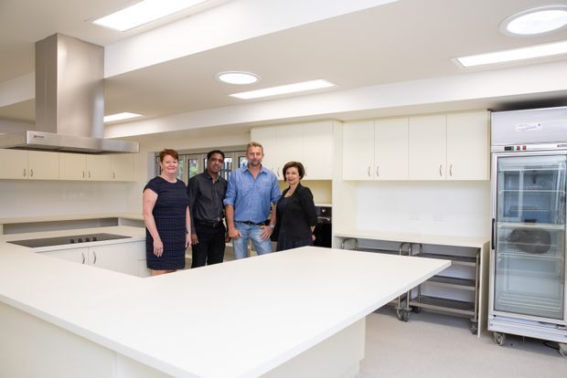 Bright, spacious commercial-style kitchen with extensive white built-in cabinetry, a large white island bench, stainless-steel rangehood, recessed and skylight-style ceiling lighting, and a tall commercial refrigerator. Four people stand near the rear work area. The room has a clean, functional, institutional fit-out with ample workspace and storage.