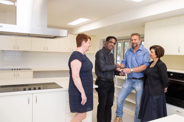 Bright, spacious white kitchen interior with extensive built-in cabinetry, a large central island with an induction cooktop, stainless-steel rangehood, integrated appliances, and large windows. Several people are standing in the kitchen, with the scene appearing to document a completed renovation or handover.