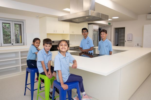 Bright, spacious commercial-style teaching kitchen with a large white island bench, extensive white cabinetry, stainless-steel rangehood, multiple stools, and several children in matching uniforms. The room has practical shelving, tiled or smooth light flooring, recessed lighting, and windows providing natural light.