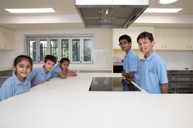 Bright, spacious contemporary teaching kitchen with a large white island, integrated black cooktop, extensive white cabinetry, stainless-steel commercial-style rangehood, tiled splashback, barred windows and several children gathered around the work surfaces.