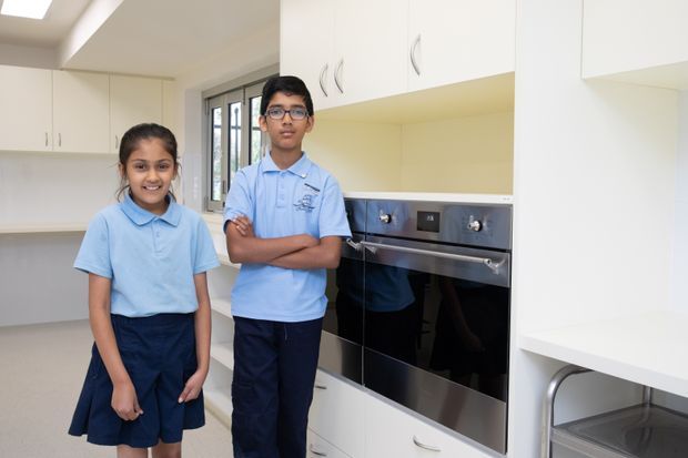 Bright, contemporary white kitchen with extensive built-in cabinetry, a wall-mounted stainless-steel oven, pale benchtops, and a skylight. Two children in school uniforms stand in front of the kitchen, while a window and partial utility area are visible in the background.