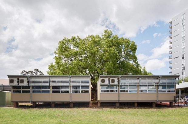 Exterior view of a low-rise modular or demountable building composed of two long wings with repetitive horizontal louvred window bands, raised above the ground on supports, and a central mature tree. A tall modern apartment or institutional building is partially visible in the background. The scene appears to be an institutional or commercial site under a bright, partly cloudy sky.