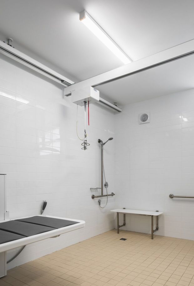 A bright, utilitarian accessible bathroom or care facility washroom with white tiled walls, beige tiled flooring, stainless-steel grab rails, a wall-mounted shower, folding wall bench, and a padded changing or treatment table. The room has a high, plain ceiling with fluorescent-style linear lighting and ceiling-mounted equipment.