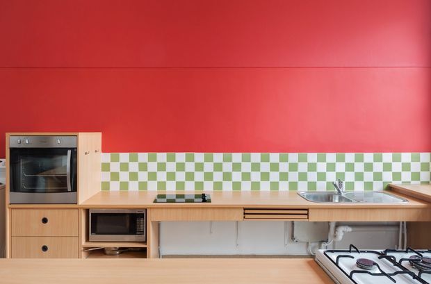 A compact contemporary kitchen with a bold red painted wall, light timber cabinetry and benchtops, and a playful green-and-white checkerboard tiled splashback. The kitchen includes a built-in oven, microwave, cooktop, double-bowl stainless-steel sink, open under-bench plumbing, and simple black hardware.