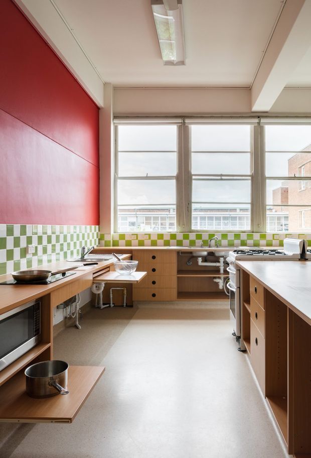 A bright, functional kitchen with extensive timber cabinetry and benchtops, a central island, exposed plumbing beneath the sink, and large multi-pane windows. The room features a bold red painted wall and a green-and-white checkerboard tile splashback, along with open shelving and integrated appliances. The space has a utilitarian, retro-inspired character with abundant natural light.