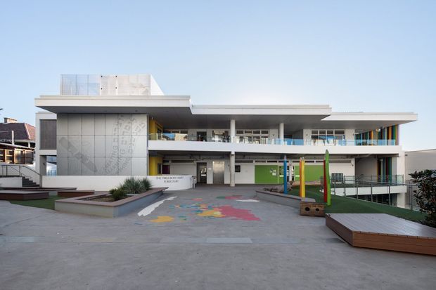Exterior view of a contemporary multi-level educational or institutional building, with broad horizontal roof slabs, extensive glazing, balconies, and a colourful paved play area in the foreground. The facade combines white rendered surfaces with darker panels and a patterned feature wall, while planted beds and timber-edged raised platforms define the forecourt.