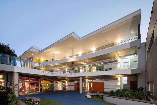 Exterior view of a contemporary multi-storey institutional or commercial building arranged around a covered courtyard. The facade features broad horizontal floor plates, extensive glazing, white rendered surfaces, exposed concrete soffits, slim metal balustrades, recessed lighting, and bold orange and yellow interior accents visible through the openings. A landscaped courtyard with blue paving and planting occupies the foreground, while signage identifies the Kleinhauer Family Building.