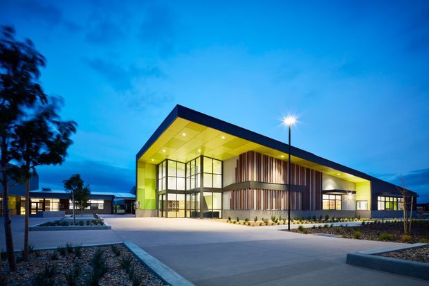 A contemporary low-rise institutional or commercial building photographed at dusk. The broad angular roof canopy has a vivid lime-yellow soffit, with extensive floor-to-ceiling glazing at the entrance, dark metal framing, and vertical timber-toned screening across the central facade. Illuminated pathways, paved forecourt areas, landscaped garden beds, and additional connected wings surround the main entrance.