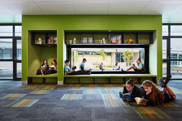 A bright contemporary library or learning commons with a vivid green built-in wall, recessed reading alcoves, open shelving, and a long window-side bench. Students are reading in the niches, on the bench, and on the patterned carpeted floor. Large windows provide views into an adjoining campus courtyard or building. The space has a suspended acoustic tile ceiling and colourful geometric carpet tiles.
