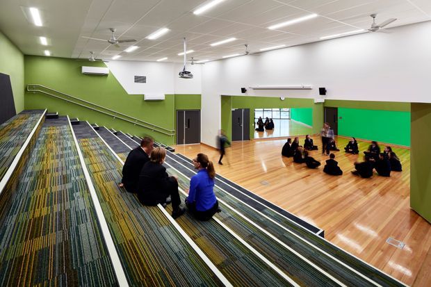 Bright contemporary multi-purpose community or school interior viewed from tiered carpeted seating. The large open room has polished timber flooring, vivid green and white walls, extensive linear ceiling lighting, multiple ceiling fans, wall-mounted equipment, and groups of people seated on the floor. A broad carpeted stepped platform with metal handrails occupies the foreground.