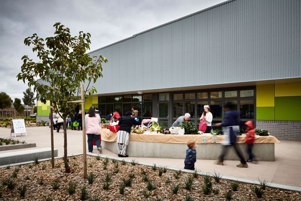 Exterior view of a contemporary community or commercial building with a large corrugated metal-clad upper facade, dark-framed glazed frontage, and bright lime-green accent panels. People gather around a long built-in concrete planter or counter displaying produce or plants, with landscaped gravel beds and young trees in the foreground.
