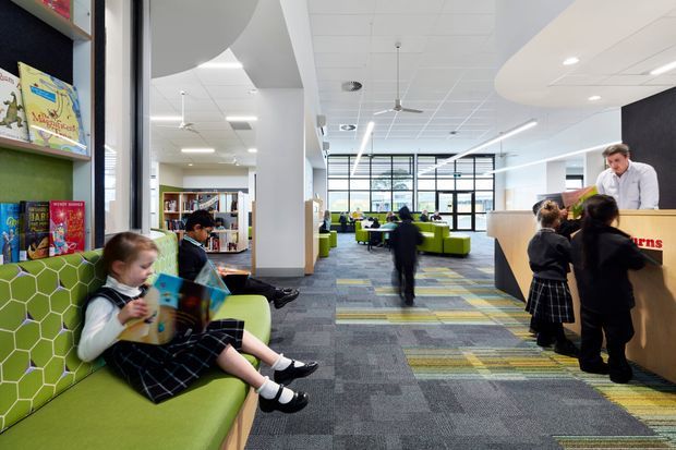 A bright contemporary school library or learning commons with children reading on a lime-green upholstered built-in bench, bookshelves, lounge seating, study tables, and a staffed timber reception or circulation desk. The open-plan space has abundant glazing, a suspended grid ceiling with linear lighting and ceiling fans, colourful patterned carpet, and a lively educational atmosphere.