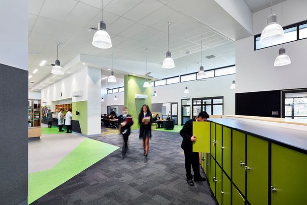 A bright, open-plan contemporary commercial interior, likely an educational or workplace common area. The space features high ceilings with suspended linear grid panels, numerous white pendant lights, clerestory-style glazing, white walls, vivid lime-green accent walls and built-in storage, dark charcoal carpet tiles, and multiple people moving through the area. A large lime-green storage or locker unit with a pale benchtop dominates the foreground, while workstations, shelving, and glazed rooms are visible in the background.
