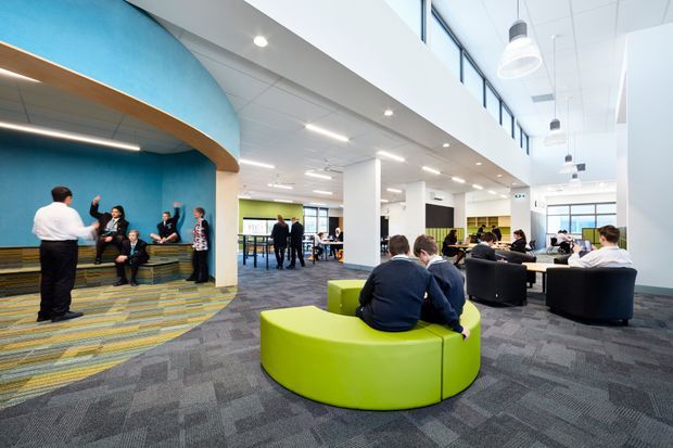 A bright, contemporary open-plan commercial interior, likely a school or workplace commons area. The large space has white structural columns, a suspended tiled ceiling with pendant and recessed lighting, high-level glazing, grey patterned carpet, and clustered lounge seating. A vivid turquoise curved feature wall and arched timber-edged alcove define a social breakout area, while lime-green circular upholstered seating forms a bold focal point in the foreground.