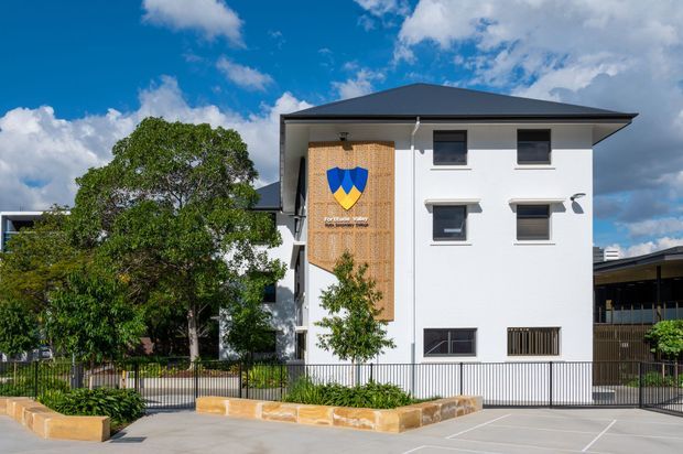 Exterior view of a contemporary multi-storey white building with a low-pitched dark hipped roof, projecting eaves, dark-framed windows, and a prominent timber-toned vertical feature panel bearing a shield emblem. The building is set behind landscaped planting, metal fencing, and paved parking areas under a bright blue sky.