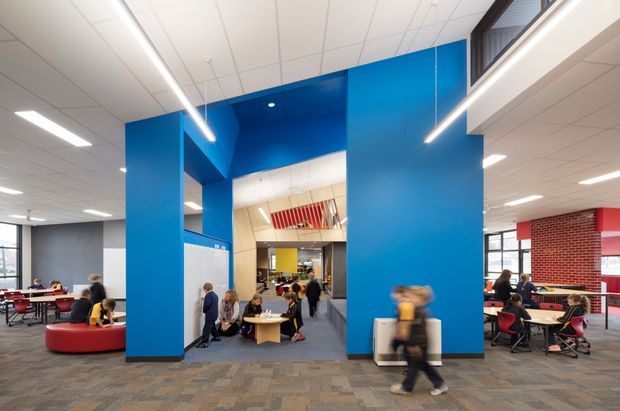 A bright contemporary commercial interior, likely an educational or institutional common area, with a large vivid blue architectural portal framing a central passage. The open-plan space includes informal lounge seating, tables and chairs, exposed red brick accents, grey carpet tile flooring, suspended linear LED lighting, and a suspended acoustic tile ceiling. People are present throughout the space, with blurred movement in the foreground.