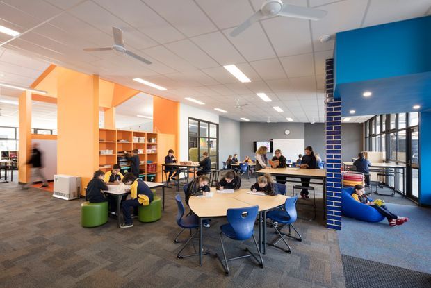 A bright, open-plan educational or library study area with students working at grouped tables, booths, and lounge seating. The space features vivid orange built-in shelving and partitions, a bold blue ceiling volume and column, exposed brick detailing, carpeted flooring, extensive glazing, suspended ceiling panels, and multiple ceiling fans and linear lights. The atmosphere is active, informal, and collaborative.