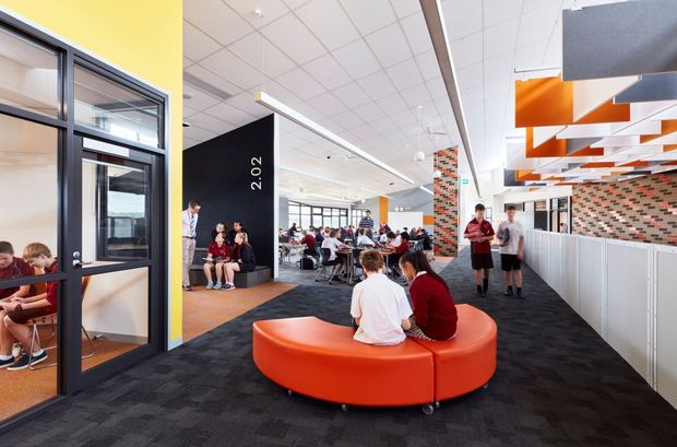 A bright contemporary school or educational common area with students seated at tables and on a large circular orange upholstered bench. The open-plan interior features black carpet flooring, white acoustic ceiling tiles, black-framed internal glazing, colourful brick-patterned feature walls, and suspended orange and white geometric ceiling panels. A yellow accent wall and high-level windows add colour and daylight. The space appears designed for informal study, gathering, and circulation.