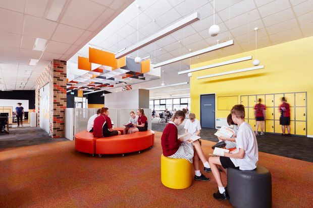 A bright, contemporary communal interior resembling a school or educational facility, with students gathered around colourful circular upholstered seating. The space features a yellow accent wall with built-in lockers, exposed brick and patterned wall surfaces, extensive suspended ceiling lighting, and a large open-plan circulation and social area. Bold orange, red, yellow, and black furnishings and ceiling panels create a lively, playful atmosphere.