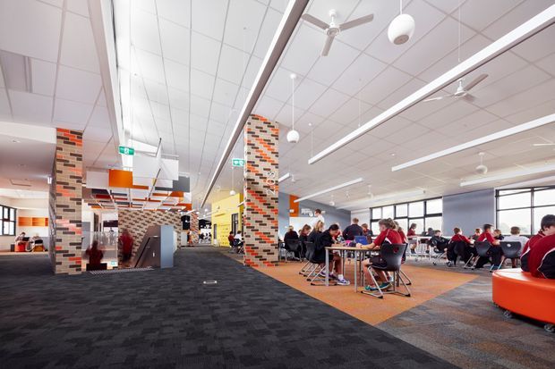 A bright, contemporary commercial dining and common area, likely in a school or institutional building. The open-plan space features extensive tables and chairs, a dark patterned carpeted circulation zone, orange carpeted dining areas, exposed brick-pattern feature columns, colourful wall sections, large windows, suspended linear lighting, ceiling fans, and modern communal furniture.