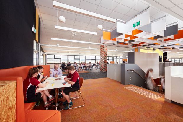 Open-plan commercial interior, likely a school or workplace common area, with students seated at tables and orange upholstered booth seating in the foreground. The space features exposed services, suspended linear lighting, spherical pendant lights, a patterned acoustic ceiling installation with white and orange panels, carpeted flooring, large windows, and a central stair or raised platform area. The palette is bright and energetic with orange, white, grey, and timber accents.